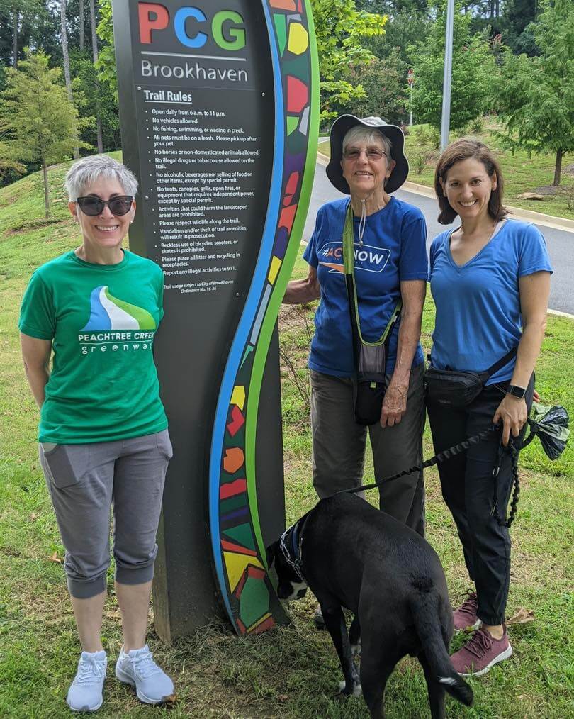 Three women and a dog standing in front of the Peachtree Creek Greenway sign